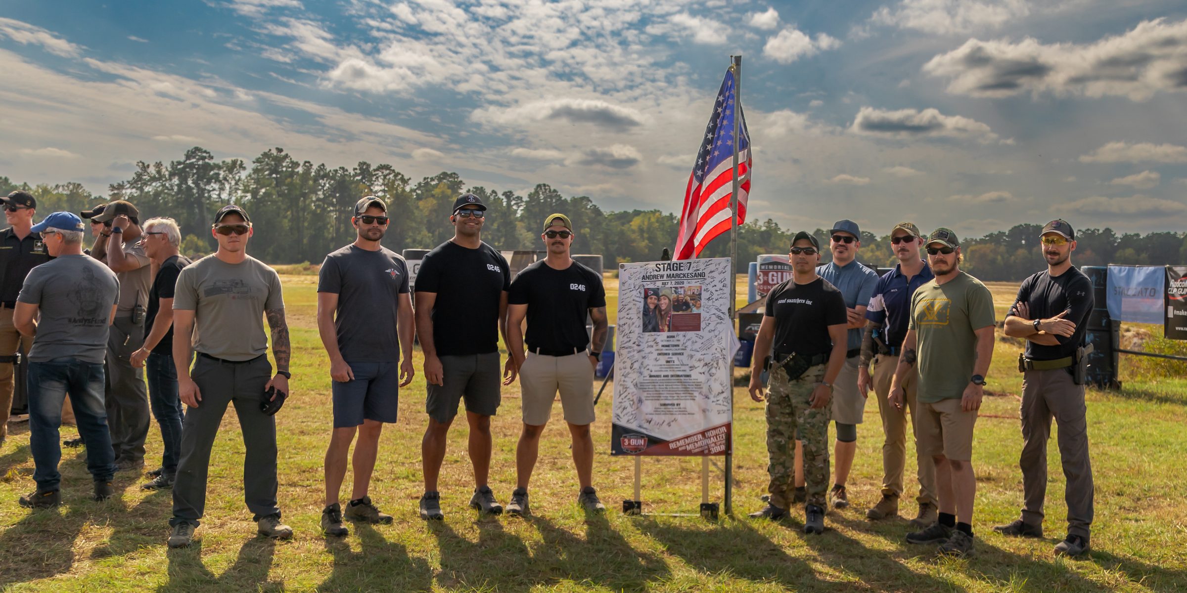 Competitors gather at an honoree stage sign and the American flag under a cloudy sky at the Memorial 3 Gun